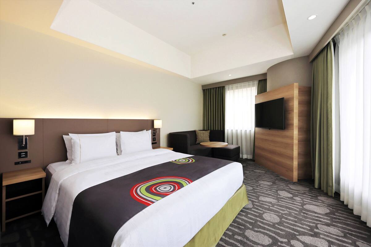 Ein Hotelzimmer mit einem großen Bett, einem Schreibtisch mit Stuhl, einem an der Wand befestigten Fernseher und einem Fenster mit Vorhängen.