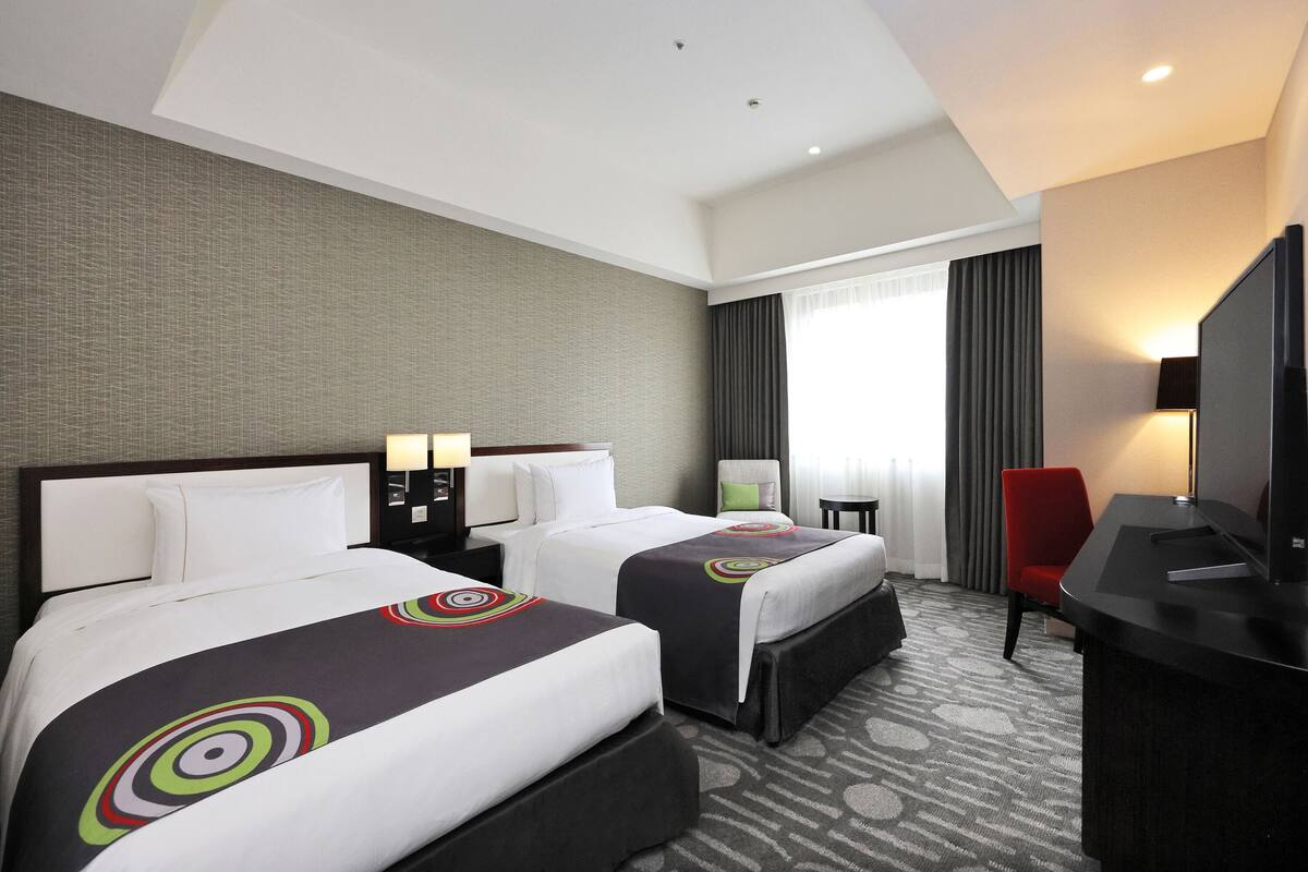Ein Hotelzimmer mit zwei Betten, einem Schreibtisch, einem Fernseher und einem Fenster mit Vorhängen.