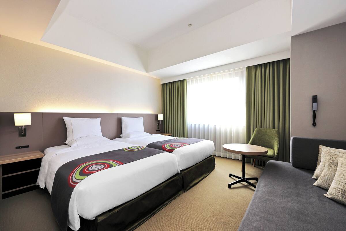 Ein Hotelzimmer mit zwei Betten, einem Schlafsofa, einem kleinen Tisch und einem Fenster mit Vorhängen.