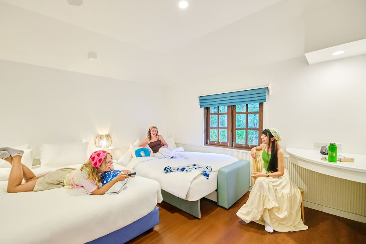 A hotel room with two beds, a window with blue curtains, a bedside table with a phone and a green bottle, and a lamp on the left side.