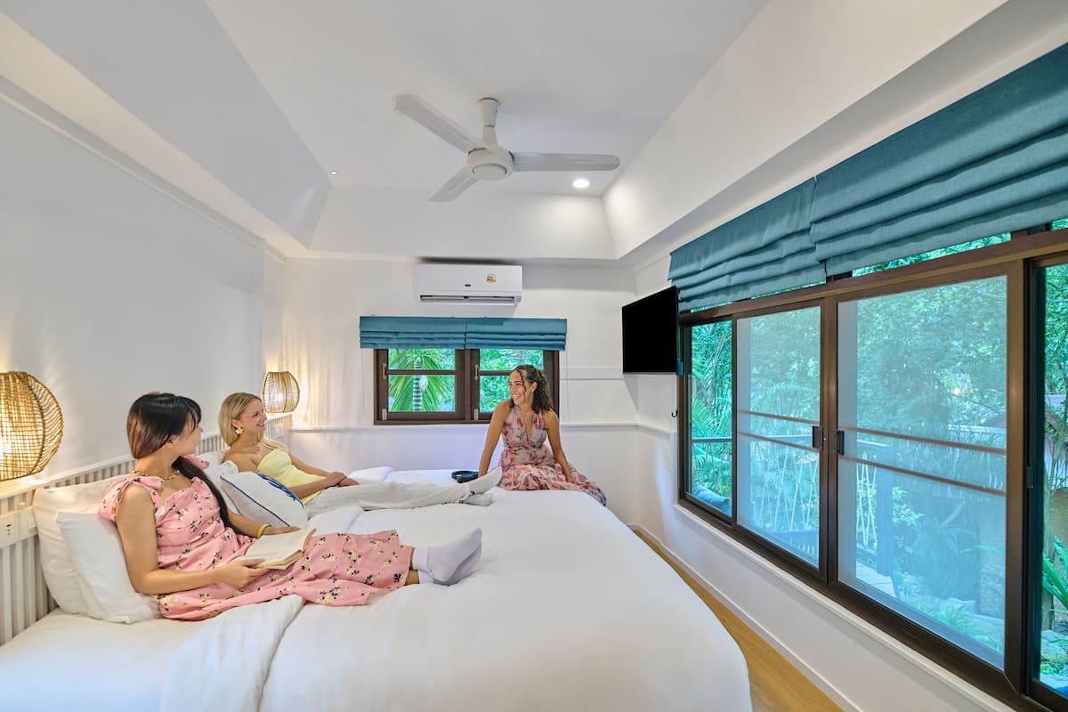 A bedroom with a large bed, a ceiling fan, an air conditioning unit, a window with curtains, and a wall-mounted TV.