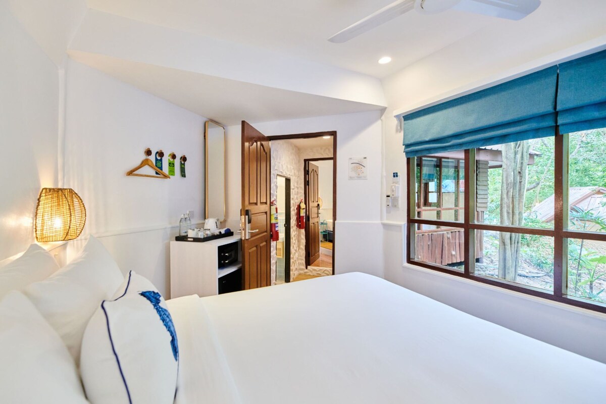 Una habitación de hotel con cama, mesita de noche, espejo, ventana con vistas y lámpara fijada a la pared.