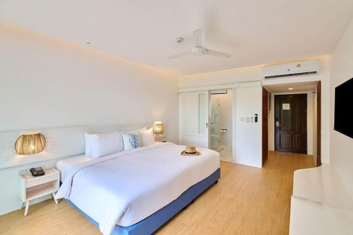 A hotel room with a bed, bedside tables, a ceiling fan, an air conditioning unit, and a door leading to a bathroom.