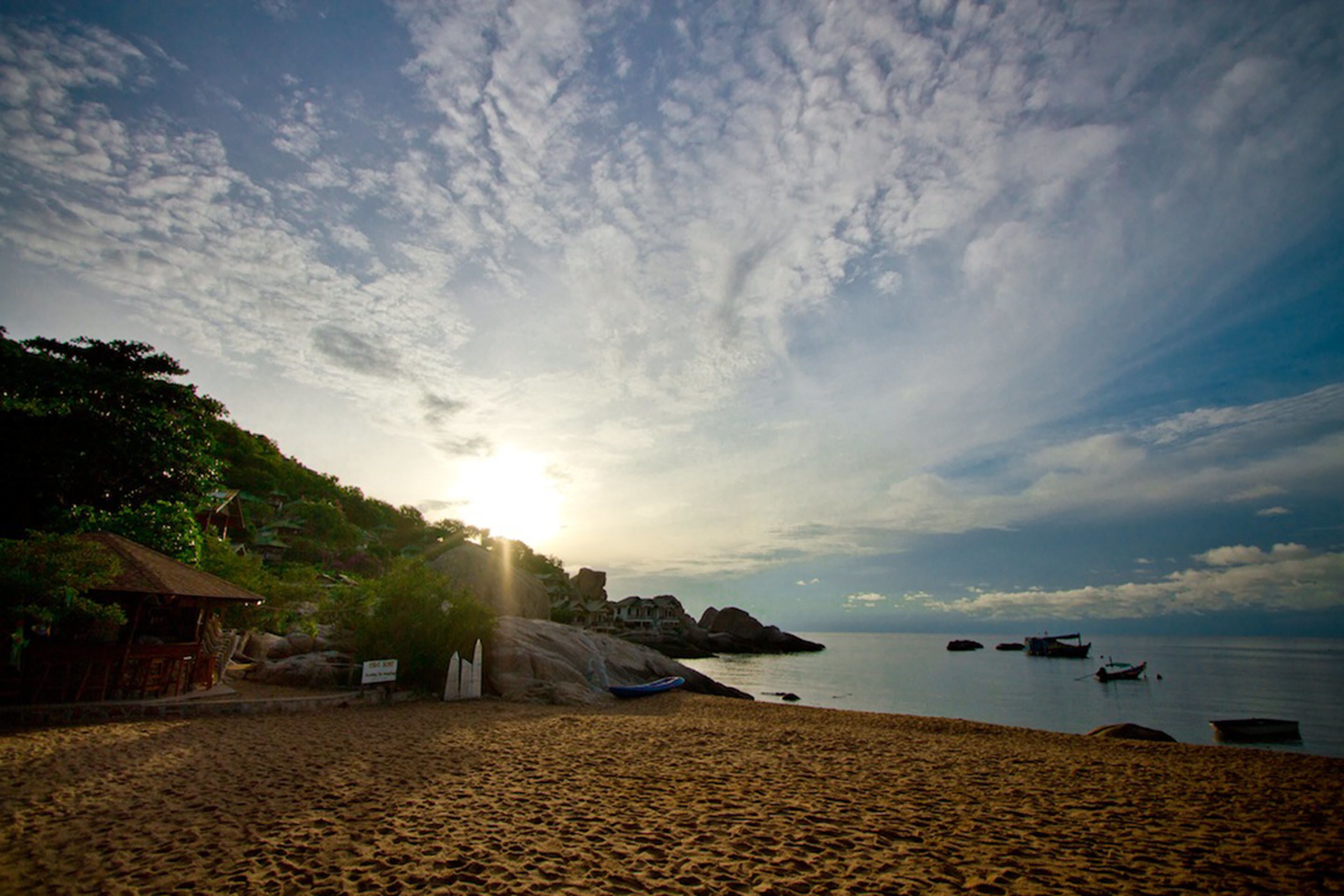 A beach with a hut, rocky outcrops, and boats in the water.