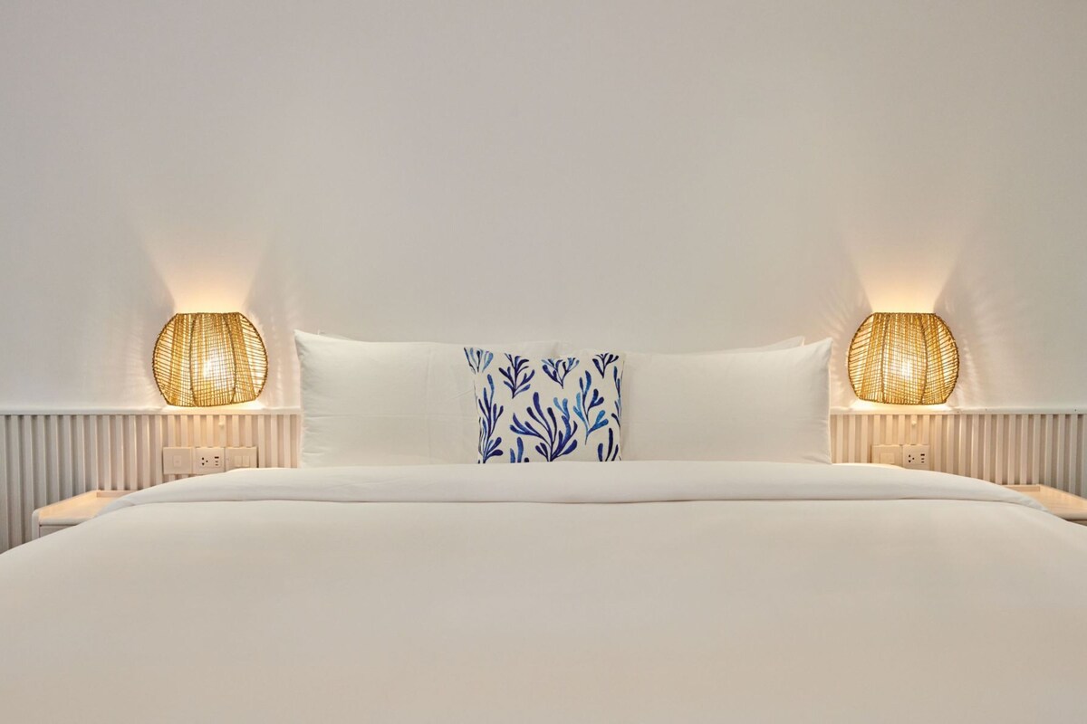A neatly made bed with white bedding, two bedside lamps with woven shades, and a single decorative pillow with a blue leaf pattern.