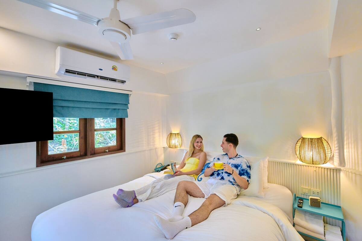 A bedroom with a bed, a ceiling fan, an air conditioning unit, a window with a view, and a bedside table with a lamp.