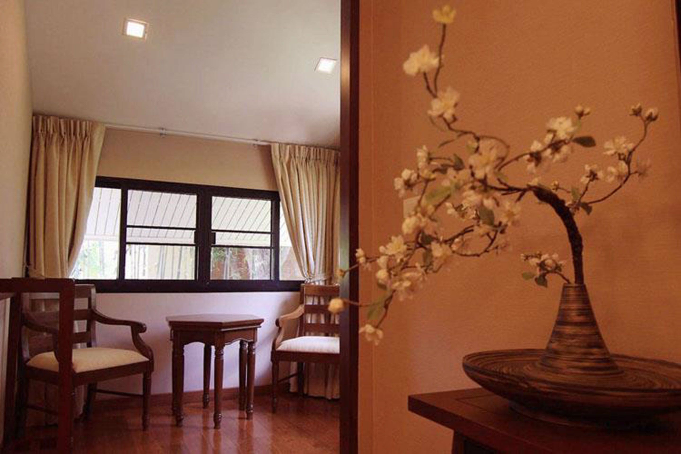 Una habitación con un ventanal grande, una silla de madera, una mesita y un jarrón con flores.