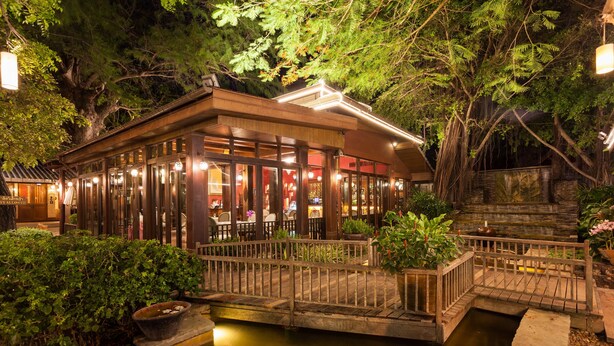 Een traditioneel Japans restaurant met een houten gevel, een kleine brug over een waterpartij en weelderige begroeiing rondom.