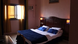 A hotel room with a bed, a desk, a chair, a window with curtains, a painting on the wall, and a wall-mounted air conditioner. - Prestige Hotel (Yaoundé)