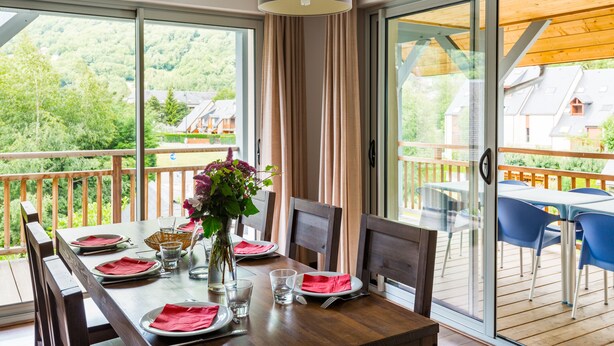 A dining area with a table set for four, a vase with flowers, and a view of a forested area through large windows. - Résidence Lagrange Vacances Le Clos Saint Hilaire (Saint-Lary-Soulan)