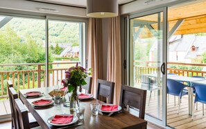 Un coin repas avec une table dressée pour quatre personnes, un vase rempli de fleurs et une vue sur une zone boisée, visible à travers de grandes fenêtres.