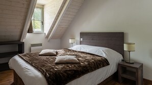 A bedroom with a sloped ceiling, a bed with a brown blanket and pillows, two bedside lamps, and a window with a view of greenery. - Résidence Lagrange Vacances Le Clos Saint Hilaire (Saint-Lary-Soulan)