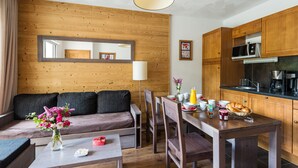 A modern kitchen with wooden cabinets, a dining table with chairs, a sofa, and a small coffee table with a vase of flowers. - Résidence Lagrange Vacances Le Clos Saint Hilaire (Saint-Lary-Soulan)