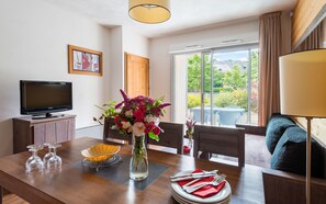 A modern dining area with a wooden table, chairs, a vase of flowers, a television, and a view of the outdoors.