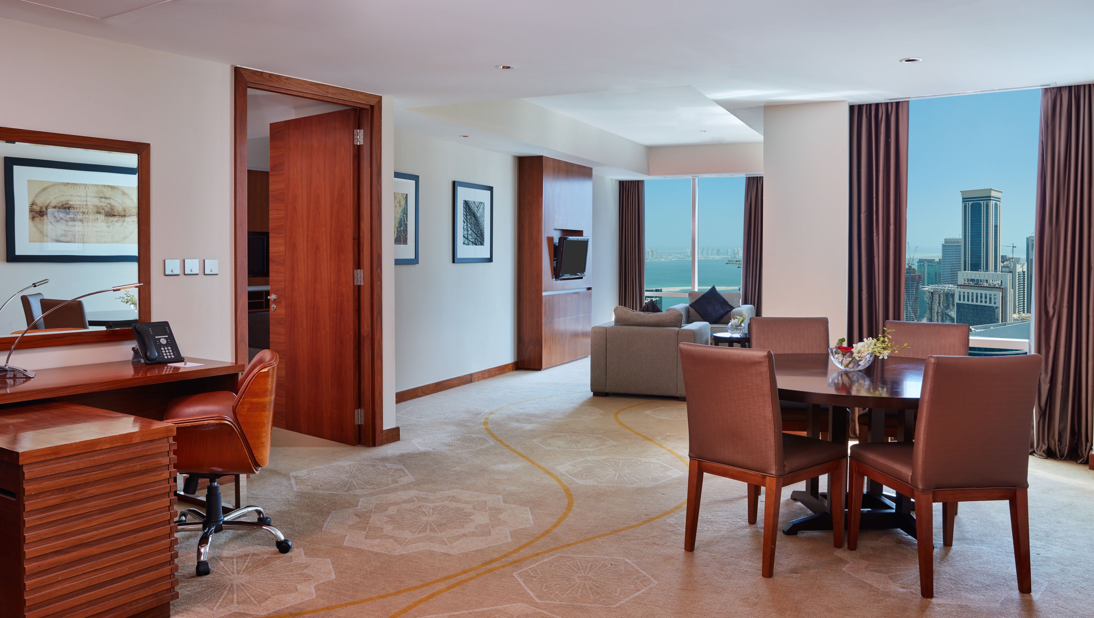 Een hotelkamer met een bureau, een bureaustoel en een zithoek met stoelen en een tafel. Er is een bank en een televisie op de achtergrond.