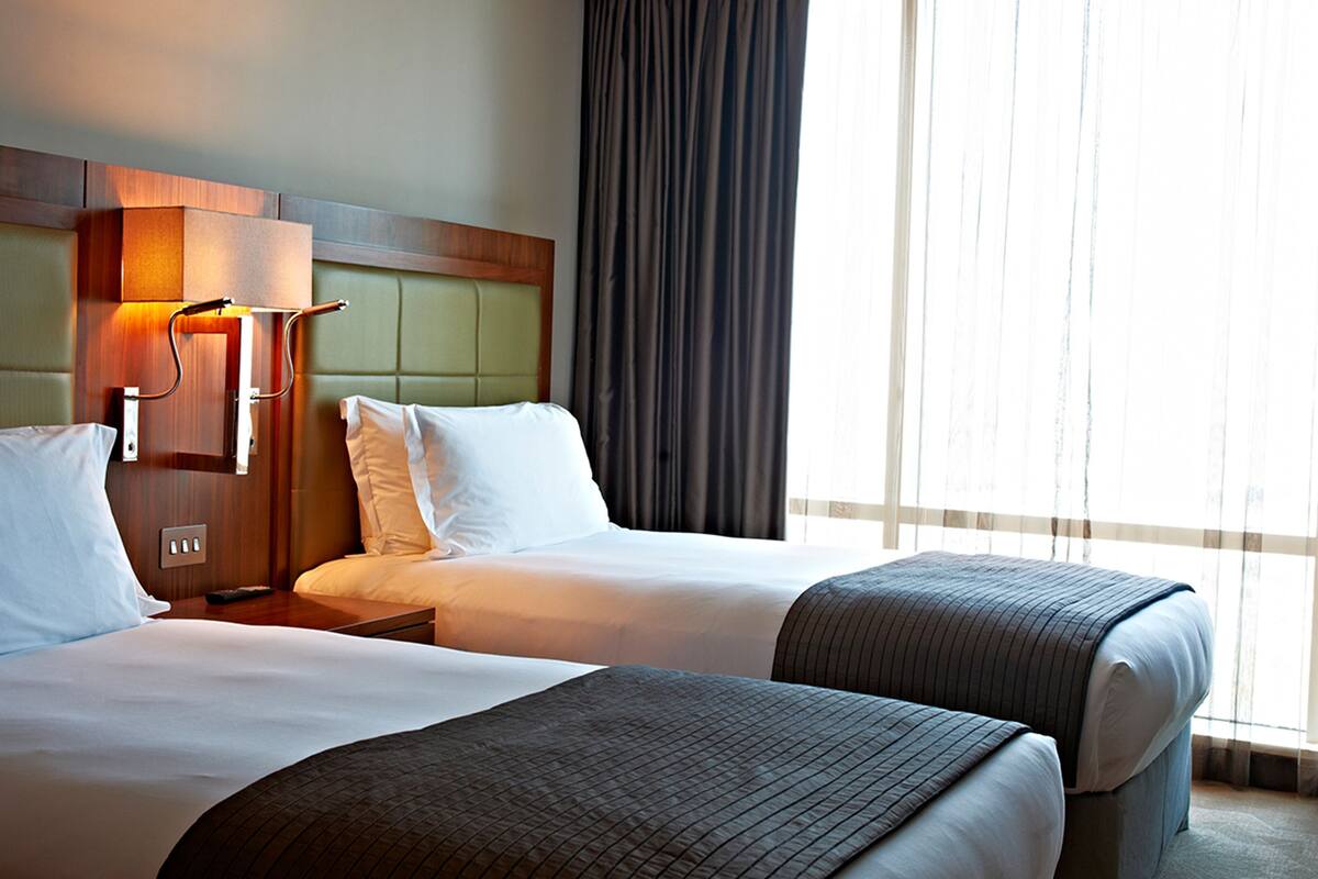 A hotel room with two beds, a wooden headboard, a nightstand, a lamp, and a large window with curtains.