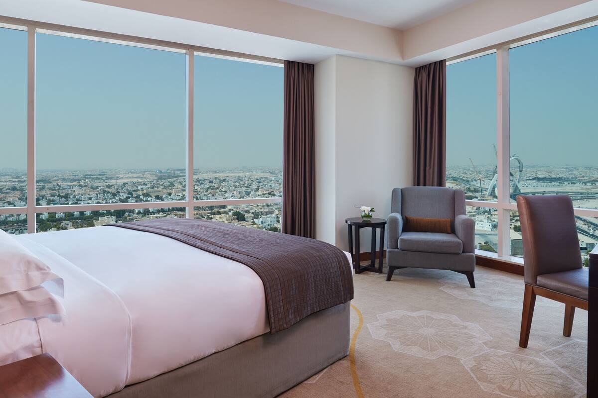 A hotel room with a large bed, a chair, a small table, and a view of a cityscape through large windows.