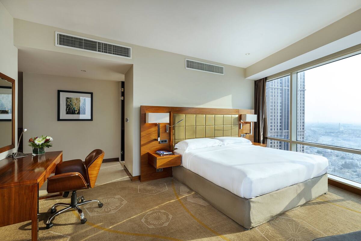 A hotel room with a large bed, a desk with a chair, a nightstand, a large window with a city view, and a framed picture on the wall.