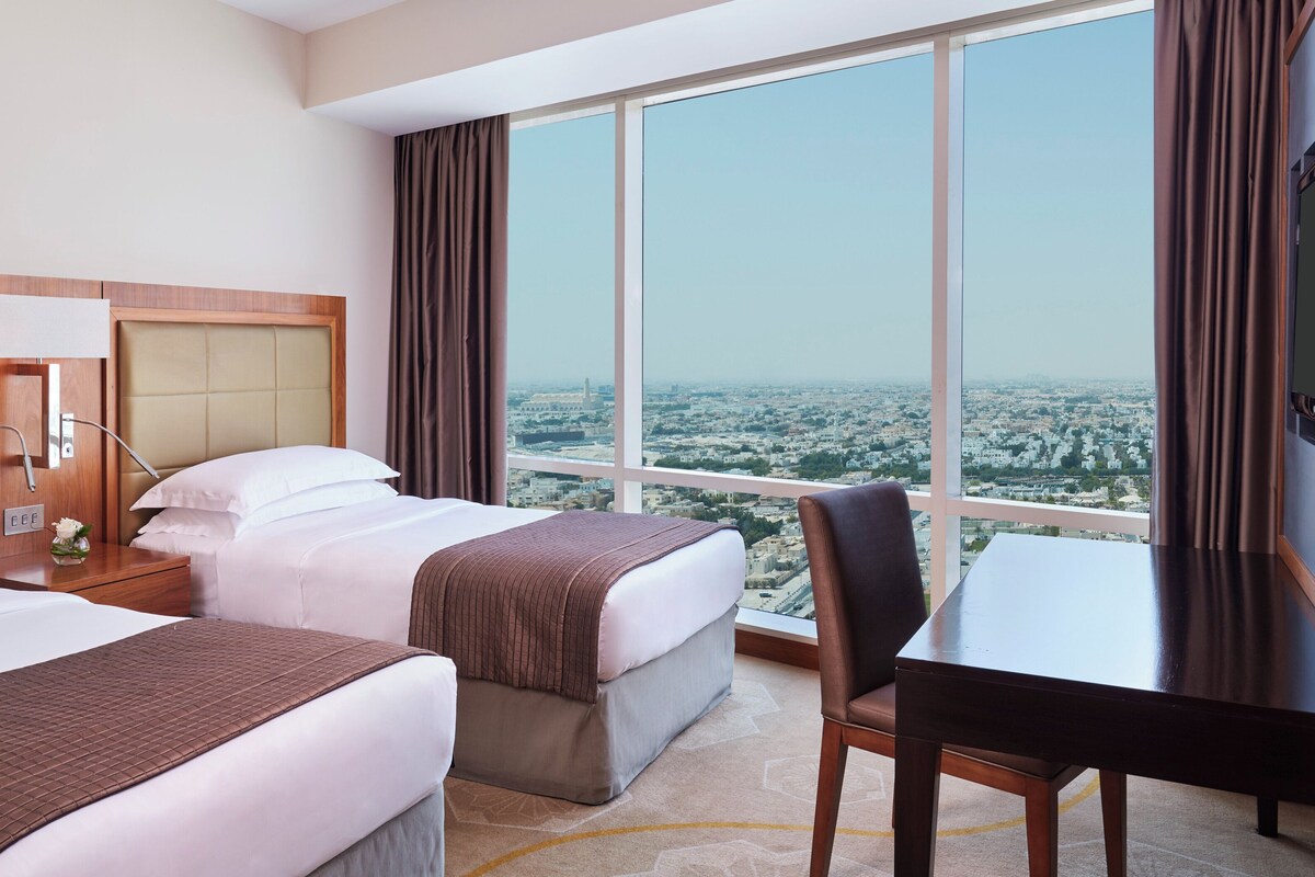 A hotel room with two beds, a desk, and a large window offering a city view.