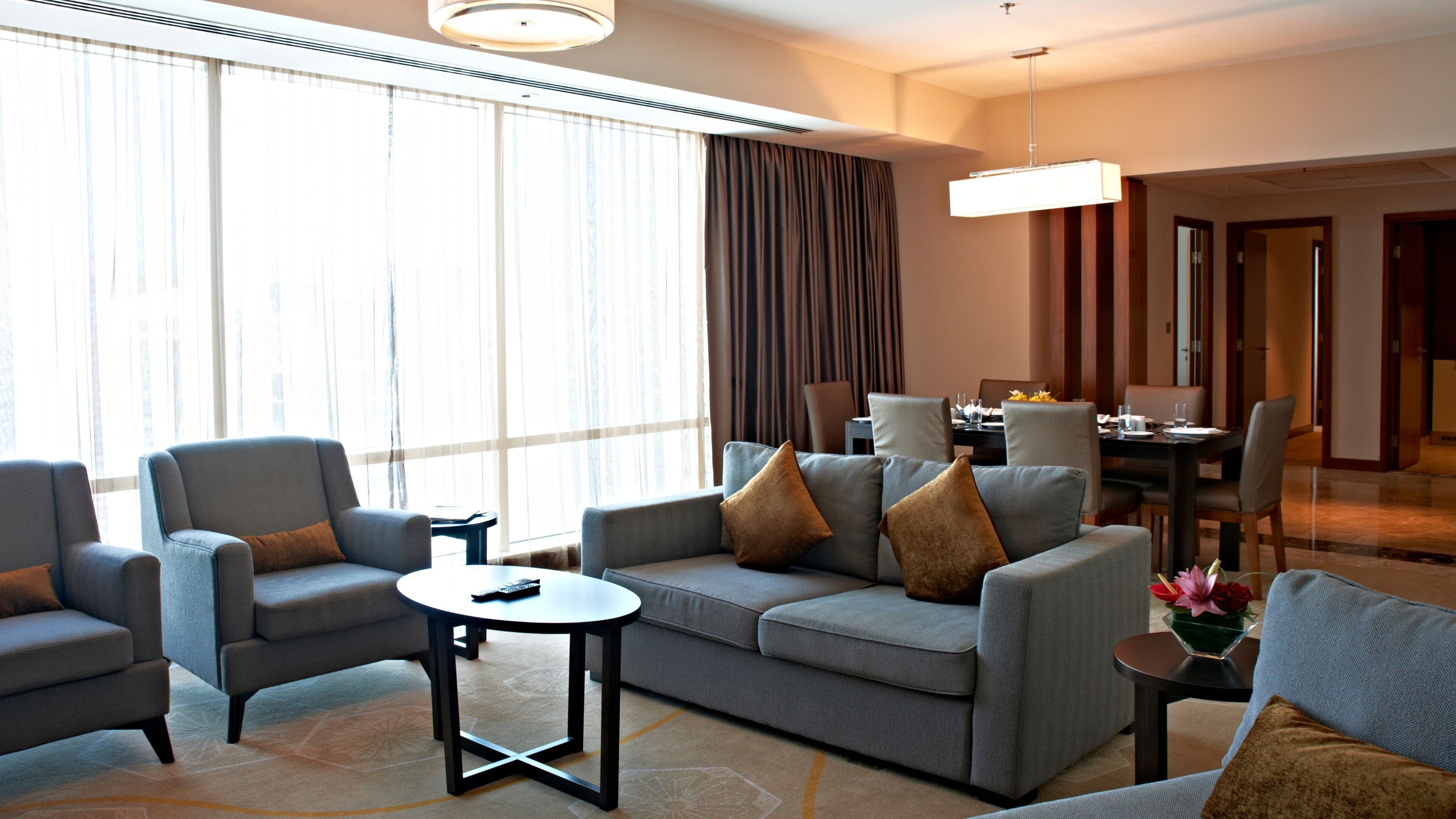 A modern hotel room with a grey sofa set, a small round table, and a dining area with a dark wooden table and chairs.
