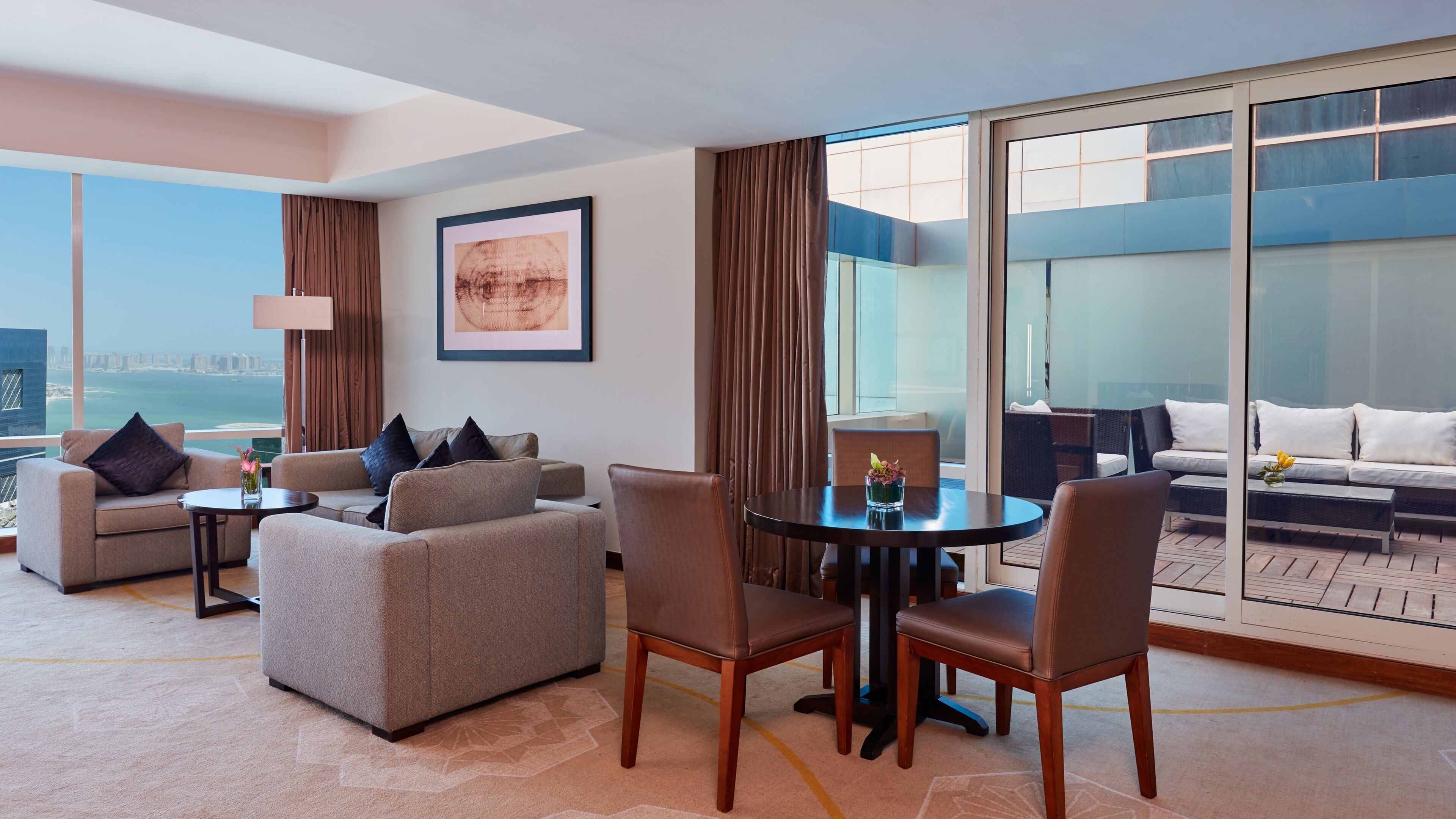 Une chambre d’hôtel avec un balcon, un ensemble de canapés, une table à manger et une grande fenêtre donnant sur la ville.