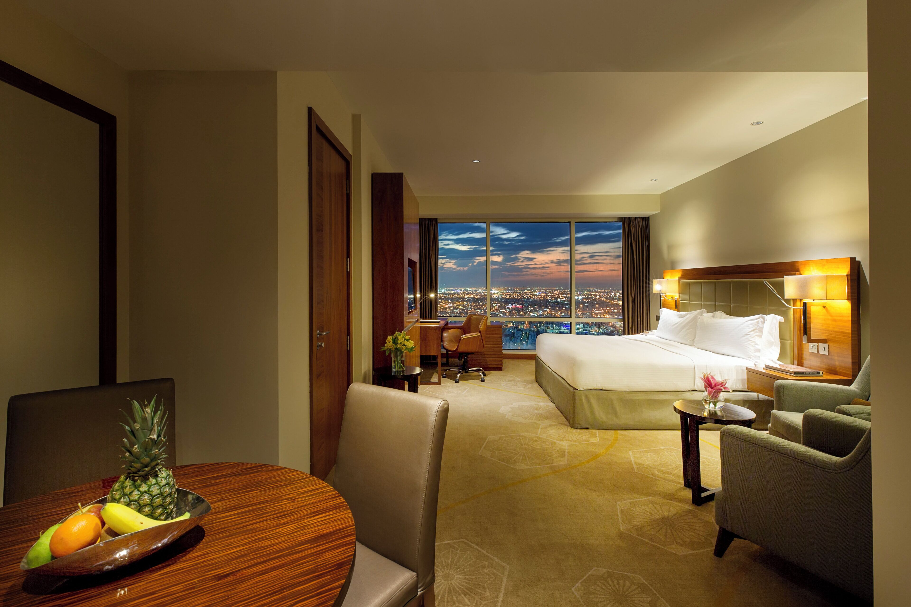 Ein Hotelzimmer mit Bett, Schreibtisch, Stuhl, einem Esstisch mit Obst und Blick auf die Stadt.