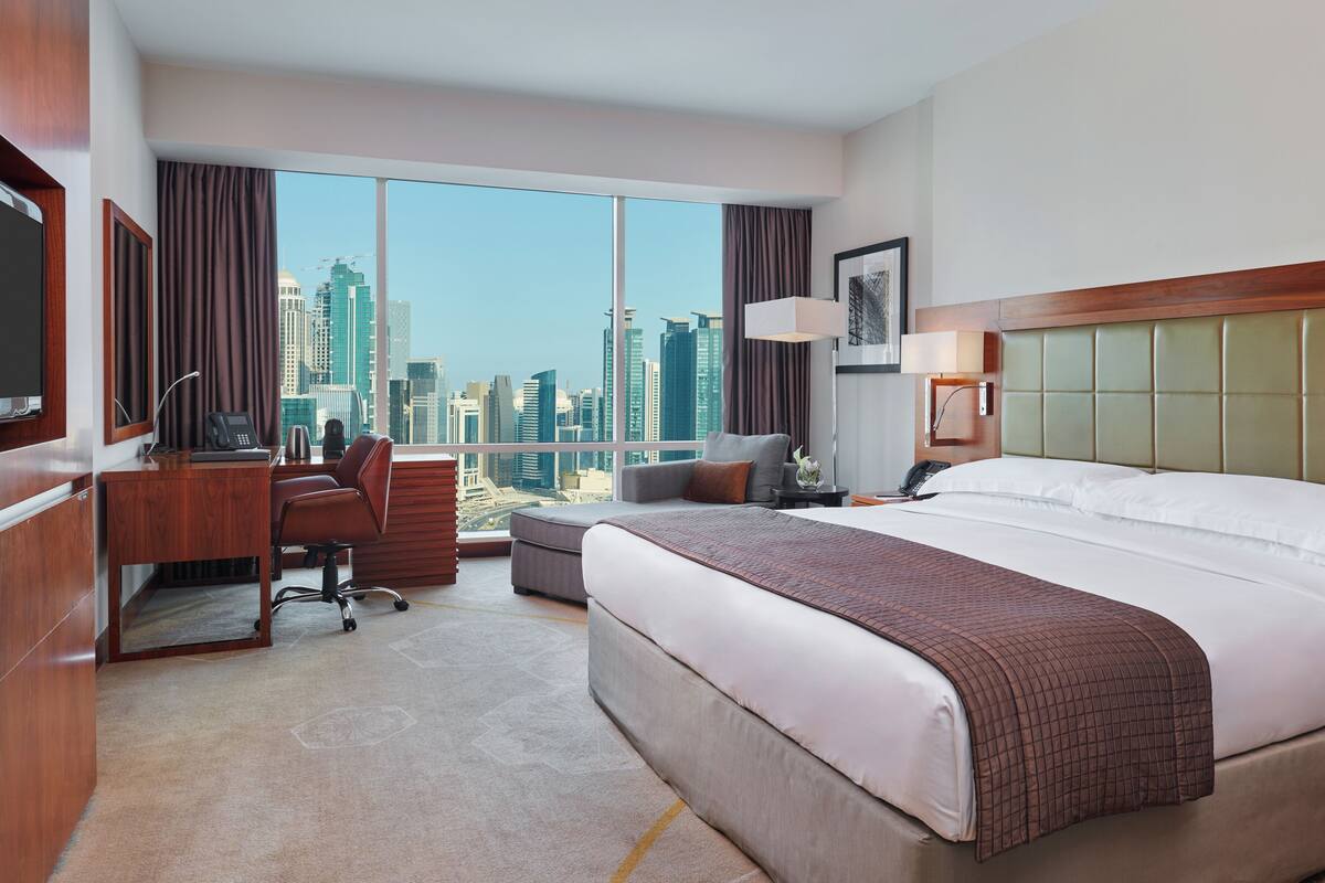 A hotel room with a large bed, a desk with a phone, a chair, a sofa, a lamp, and a view of the city skyline.
