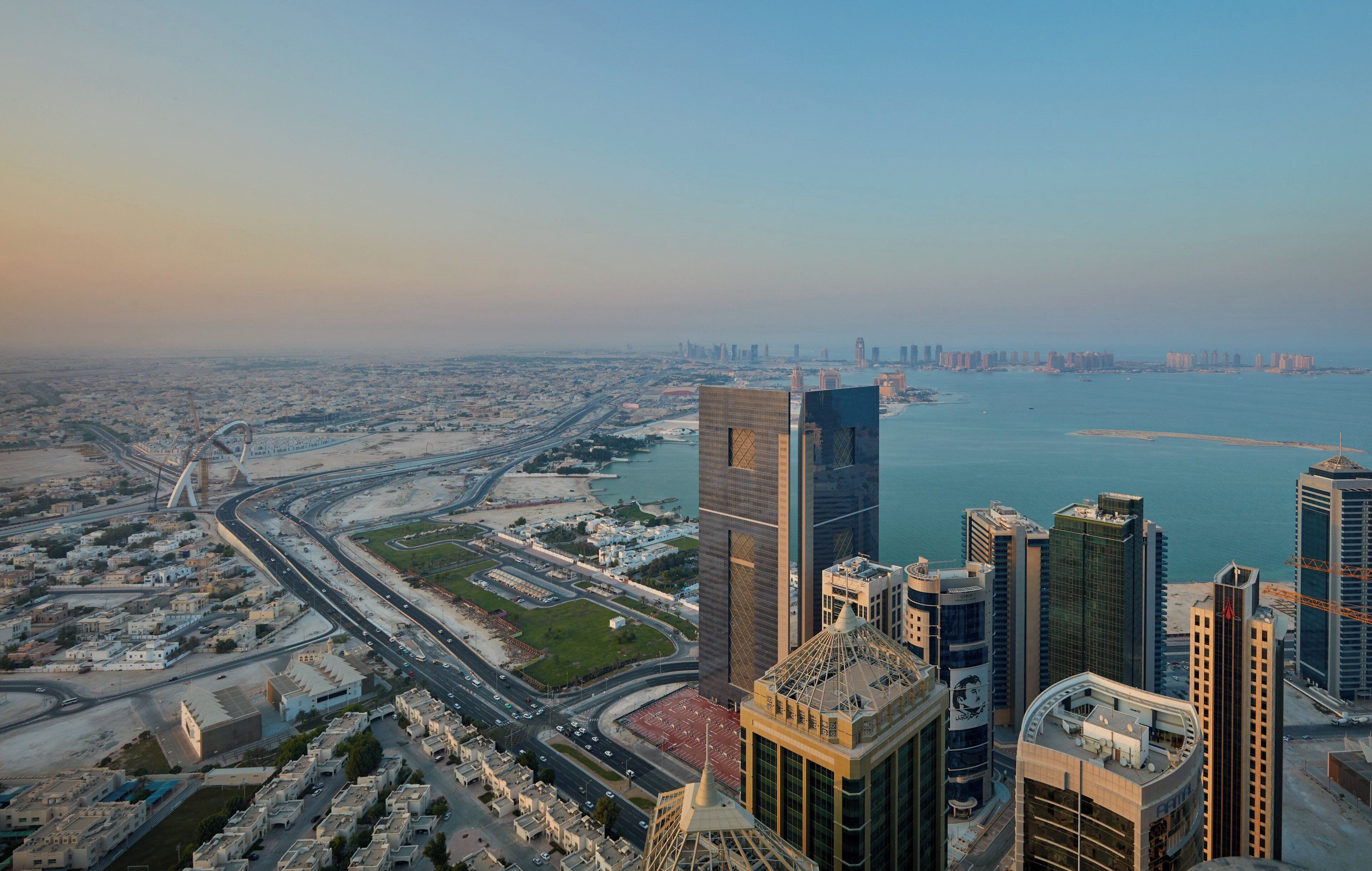 A high-rise building with a modern architectural design, situated near a coastal area with a view of the sea and a bridge in the background.