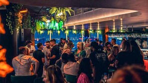 A crowded bar with people socializing, hanging plants, and a city view.