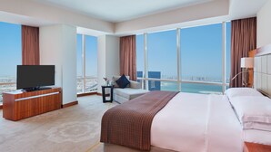 A modern hotel room with a large bed, a TV on a wooden cabinet, a small table with a vase, and a view of the city and sea through large windows. - InterContinental Doha The City by IHG (Doha)