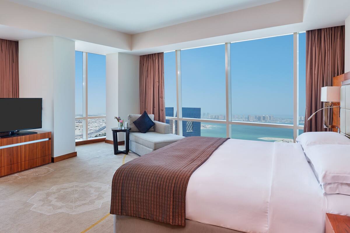A modern hotel room with a large bed, a TV on a wooden cabinet, a small table with a vase, and a view of the city and sea through large windows.