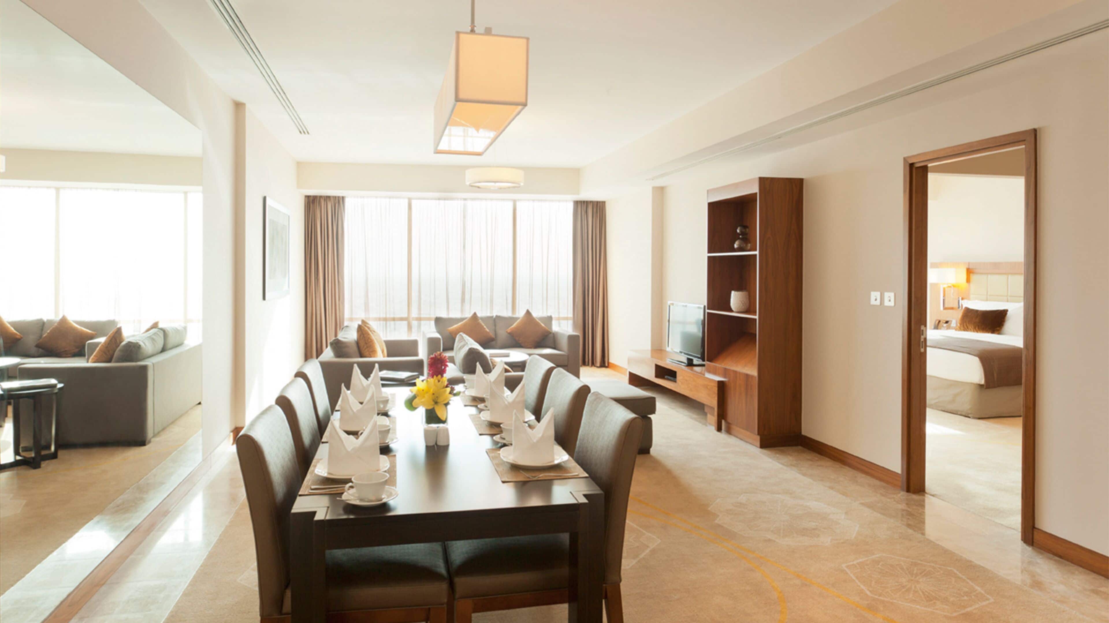 A modern hotel room with a dining area, a TV, and a bedroom visible through an open door.