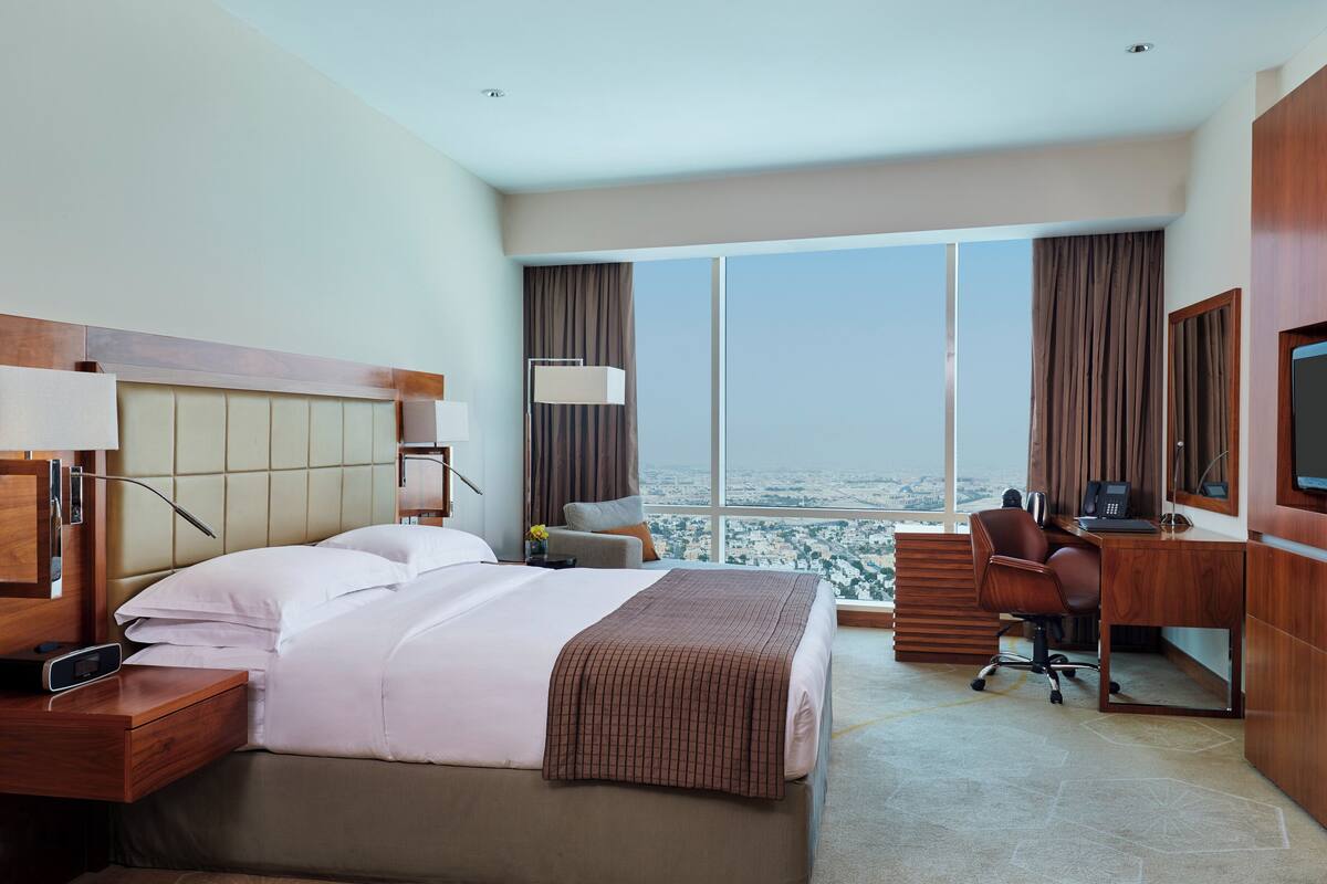 A hotel room with a large bed, a desk, a chair, a TV, and a view of the city.