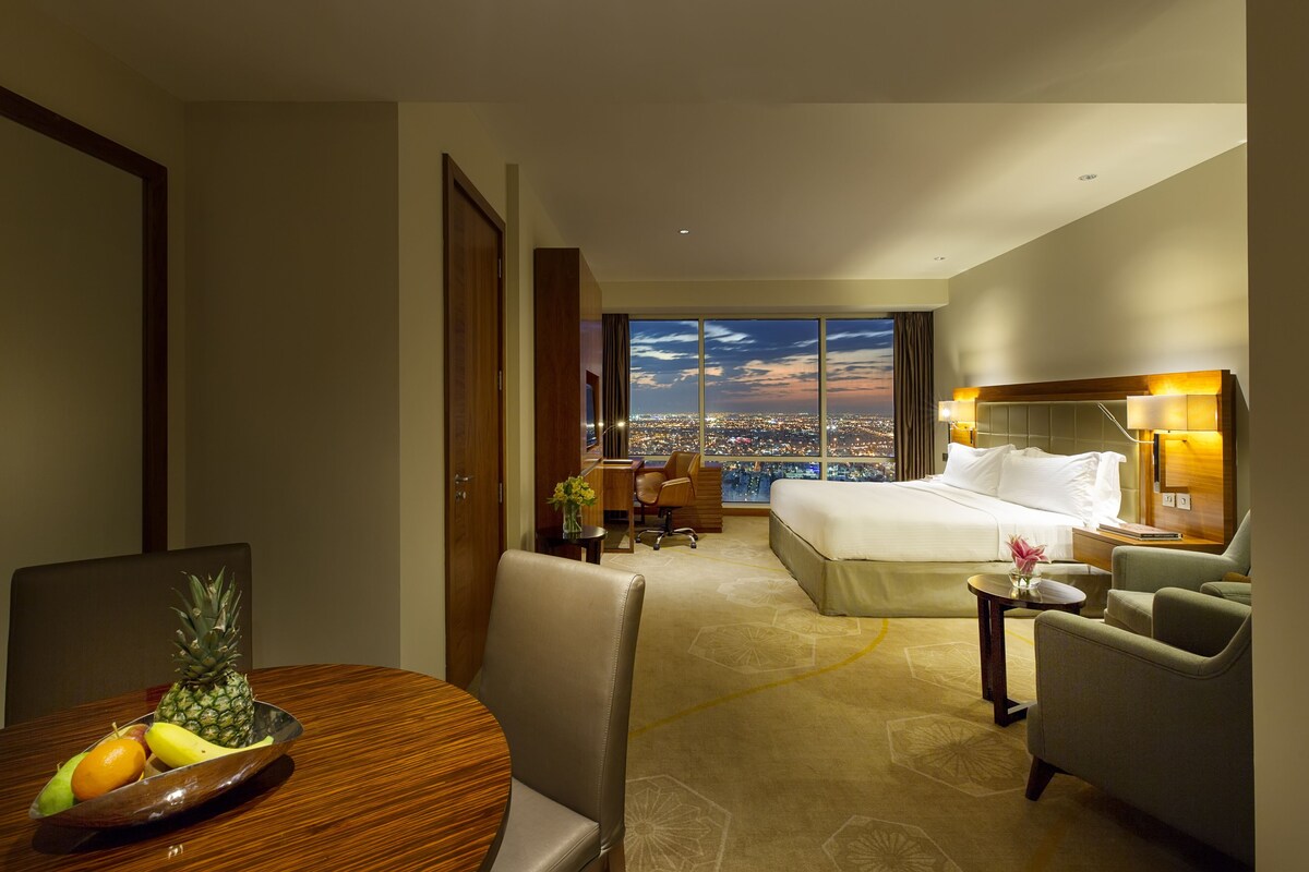 A hotel room with a bed, a desk, a dining table with fruit, and a view of the city at dusk.