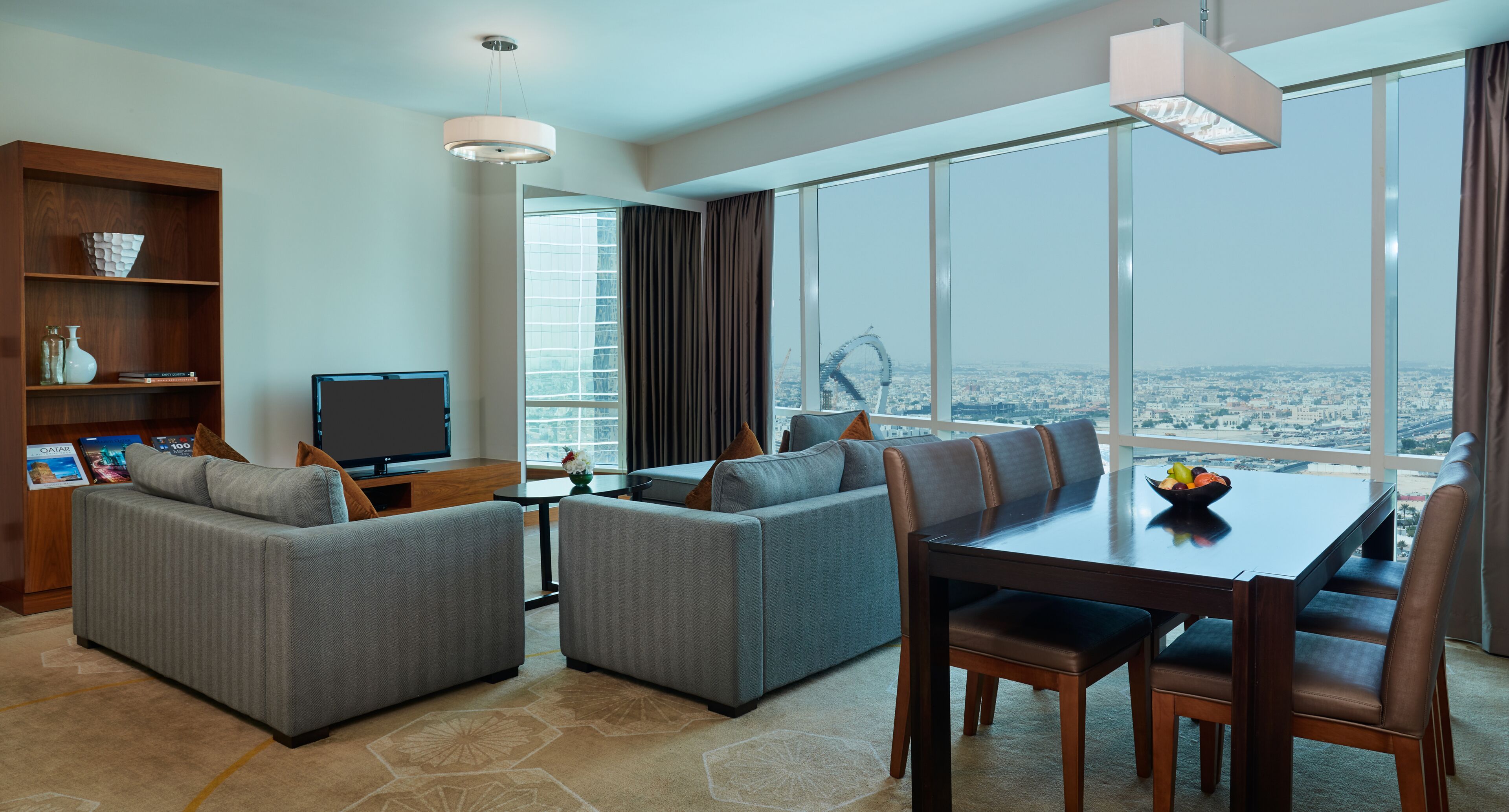 A modern hotel room with a large window offering a city view, a dining area with a table and chairs, a sofa set, and a television.