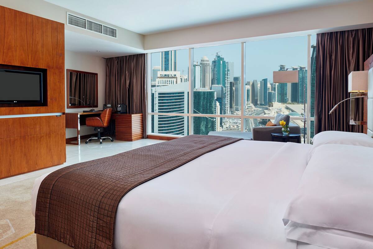A hotel room with a large bed, a desk, a chair, a TV, and a view of the cityscape.