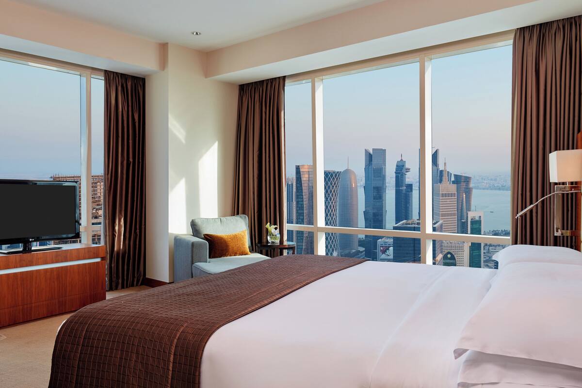 A hotel room with a large bed, a TV, a chair, and a view of a city skyline.