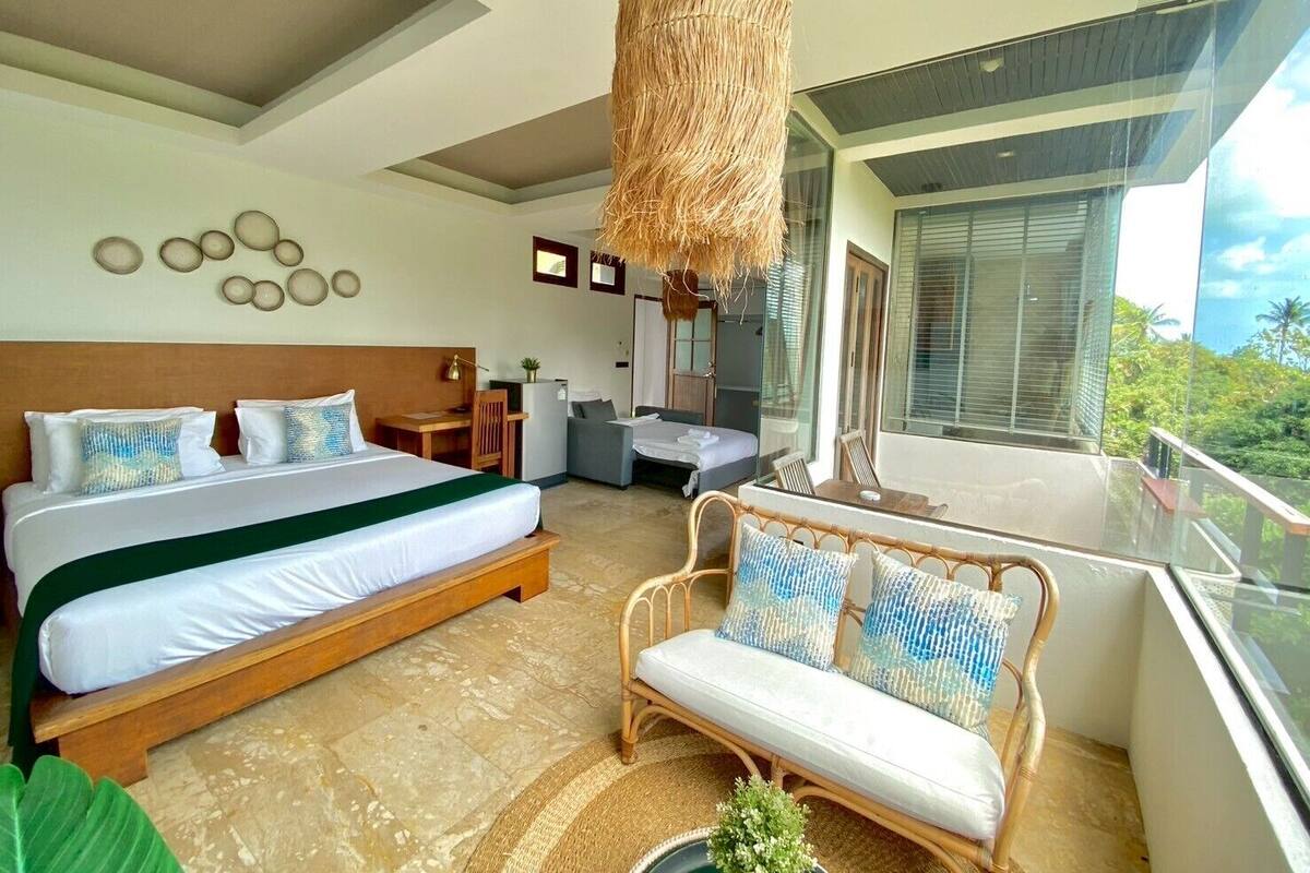A modern hotel room with a large bed, a sofa, a dining area, and a balcony with a view.