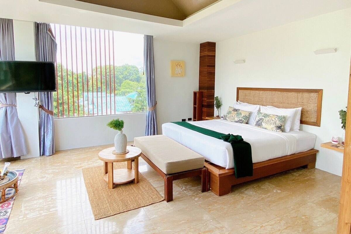 A modern bedroom with a large bed, a TV mounted on the wall, and a view of greenery outside.