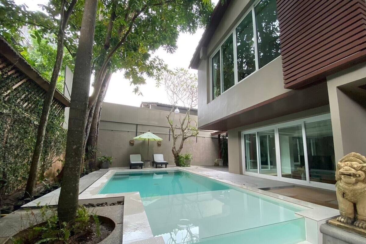A swimming pool with two lounge chairs and a small umbrella, surrounded by trees and a modern building with large windows.