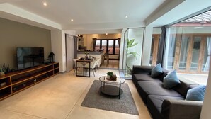 Een moderne woonkamer met een bank, een salontafel en een televisie. Er is een eethoek met een tafel en stoelen. De kamer heeft grote ramen en uitzicht op buiten.