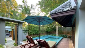 Een zwembadterras met twee ligbedden en een grote parasol, omringd door weelderige beplanting en een betegeld zwembad.