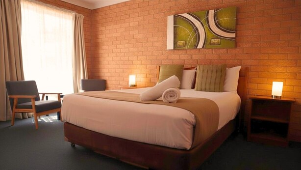 Ein Hotelzimmer mit Backsteinwand, einem großen Bett mit Holzrahmen, zwei Nachttischen mit Lampen, einem Sessel und einem Fenster mit Vorhängen.