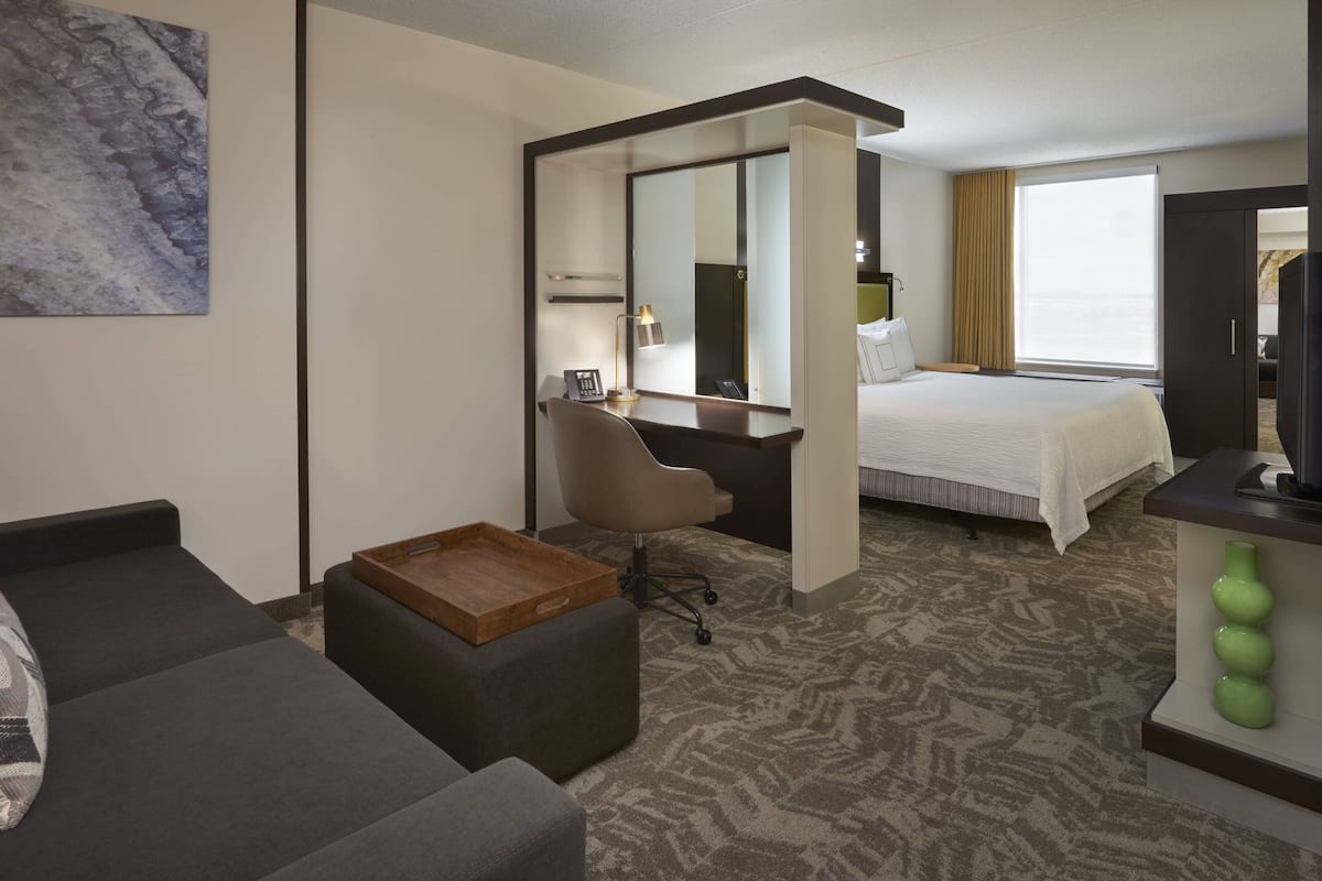 A hotel room with a bed, a desk with a chair, a small wooden table, a large mirror, and a television.