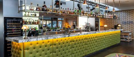 A bar area with a green cushioned bar counter, multiple shelves of liquor bottles, and pendant lighting.