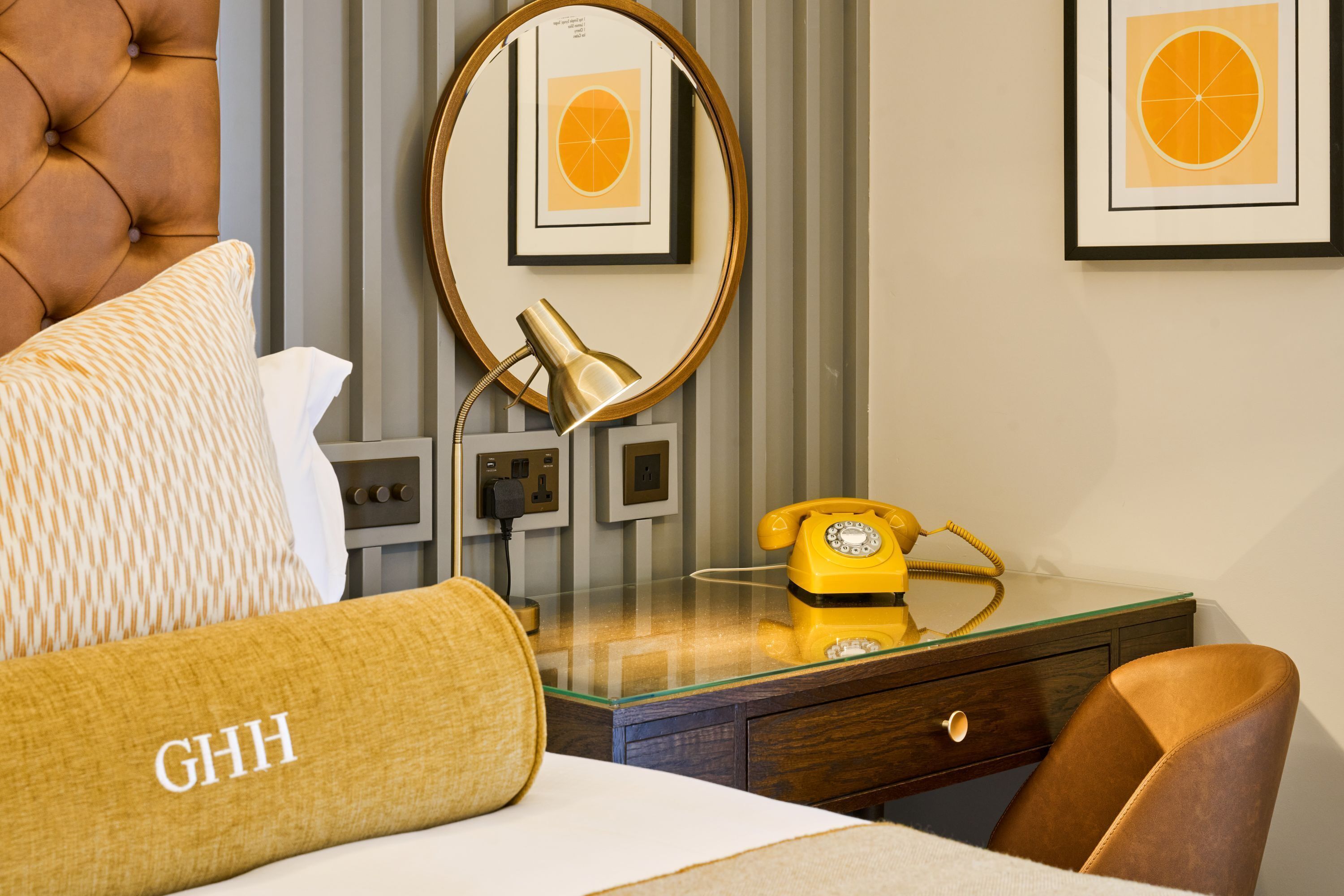 A hotel room with a tufted headboard, a bed with a patterned pillow, a glass desk with a yellow rotary phone, a mirror, and framed artwork on the wall.