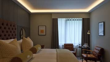 Una habitación de hotel con una cama grande, cabecera, escritorio con silla, ventana con cortinas y un cuadro en la pared.