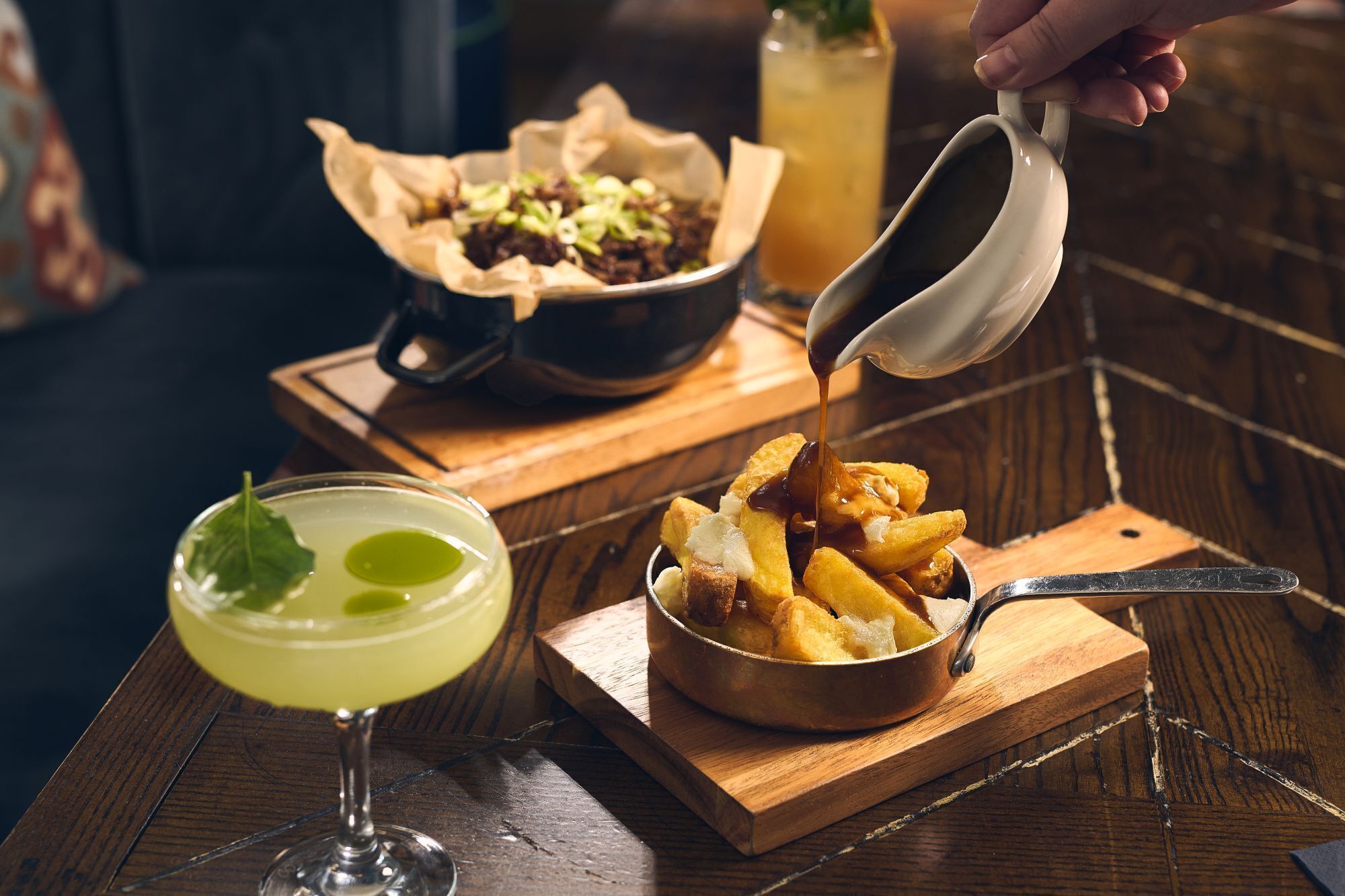 Een houten tafel met een schaal friet, een cocktail met een limoenpartje en een blikje fris.