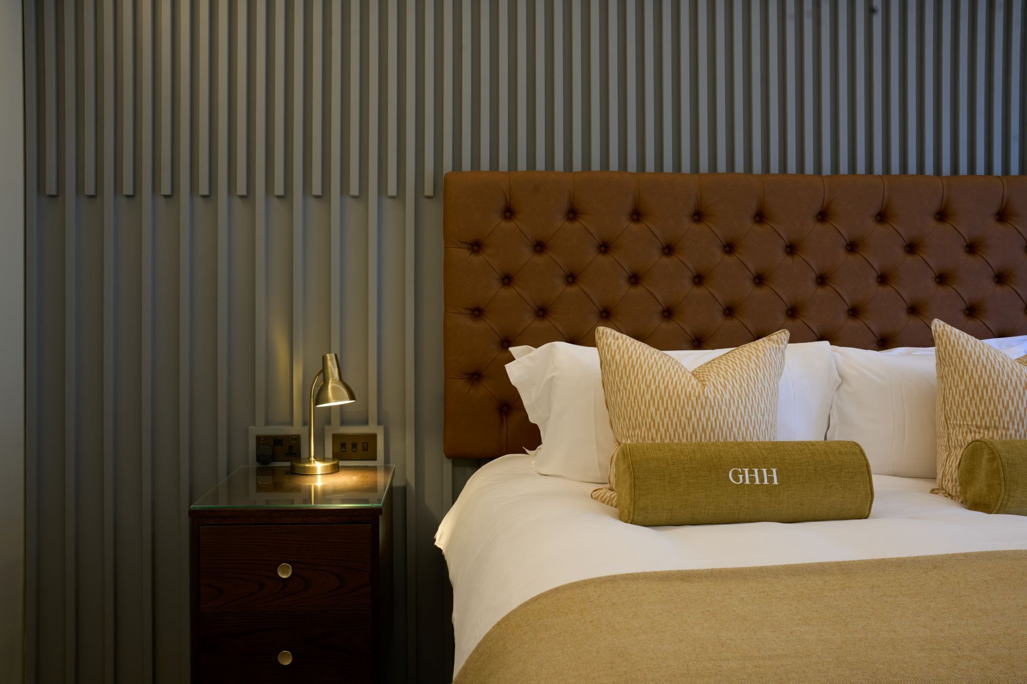 Una habitación de hotel con cabecera acolchada, una mesita de noche con lámpara, y una cama con ropa blanca y almohadas estampadas.