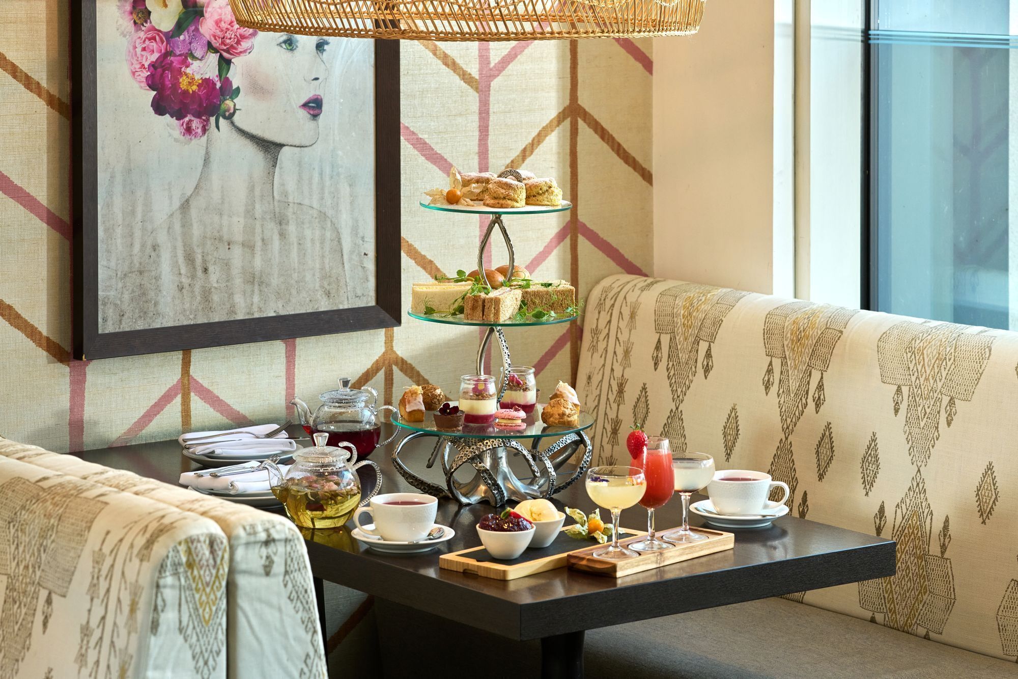 A dining area with a tiered dessert stand, a teapot, cups, and a glass of red beverage on a table.
